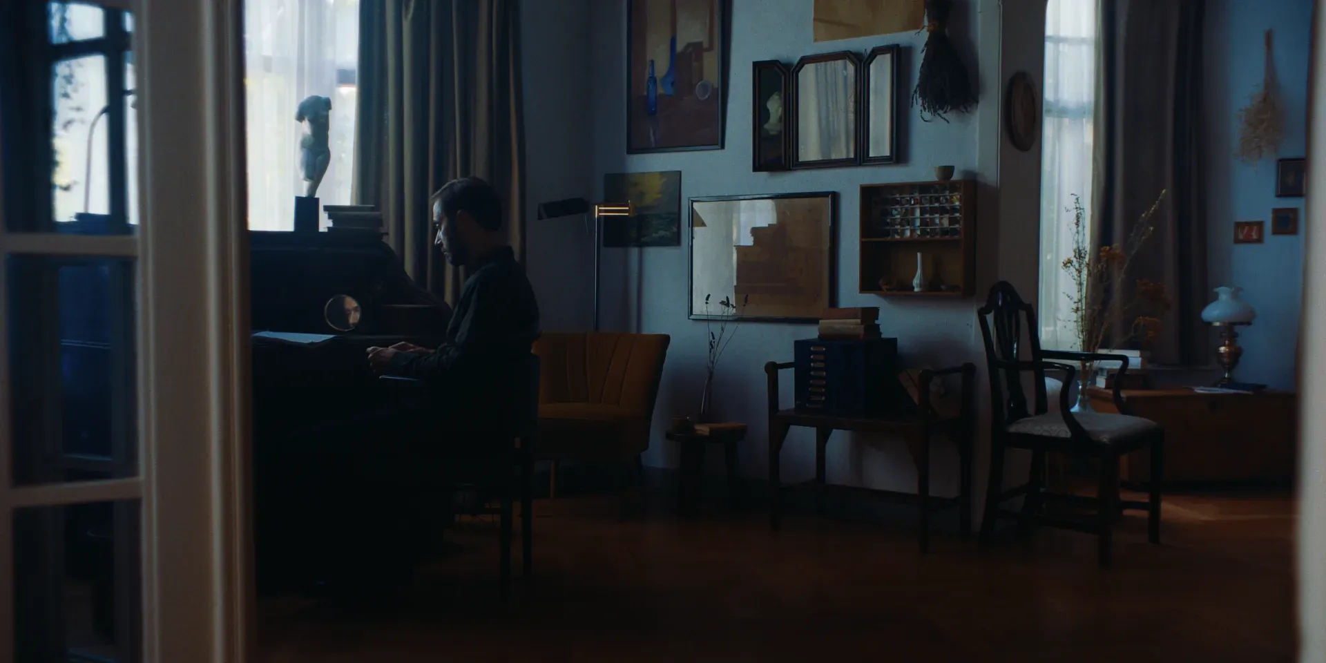 Ana's husband seated at a piano in an art-filled room in The Tears of Things